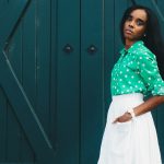 woman in white skirt and green dress shirt hand on pocket standing at the front of green door